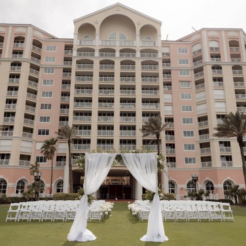 A hotel with a large wedding setup, featuring white chairs and an arch with fabric and flowers, on a lawn in front of the building.