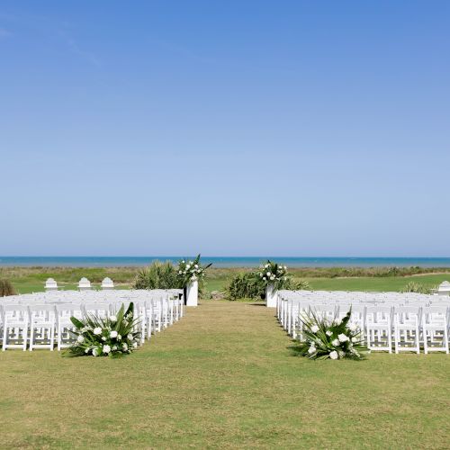 Outdoor wedding setup with white chairs and floral arrangements on a lawn by the ocean.