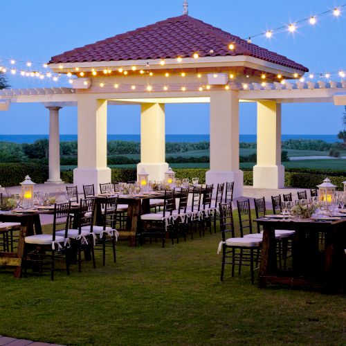 Outdoor wedding setup with string lights, tables with chairs, and a gazebo in a scenic garden with palm trees and ocean view.