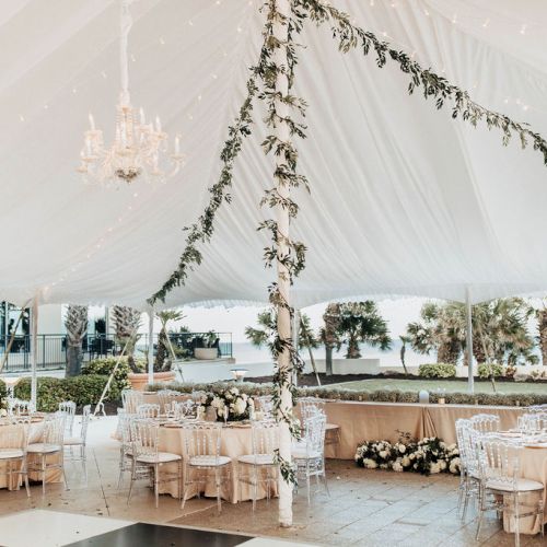 An outdoor event setup under a white tent with chandeliers, floral arrangements, and elegantly set round tables with transparent chairs.