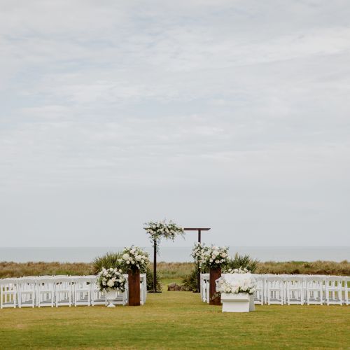 Outdoor wedding setup with white chairs and floral arrangements, overlooking the ocean. Sky is overcast.