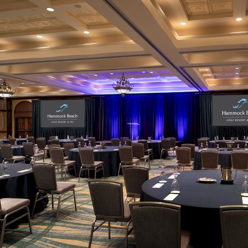 A conference room set up with round tables, chairs, glasses, name cards, and a stage with drapes and two screens displaying "Hammock Beach.”