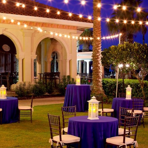 Outdoor evening event setup with round tables covered in purple cloth, surrounded by chairs, and illuminated lanterns under string lights.