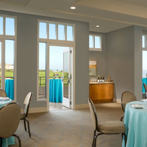 Elegant event room with round tables, blue tablecloths, wine bottles, and an ocean view through large windows.