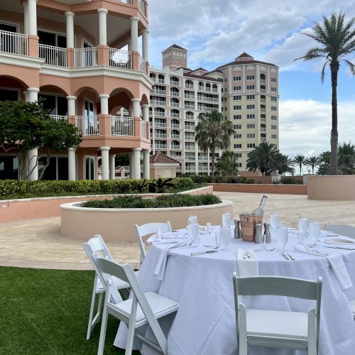 An outdoor dining setup with a table and chairs, set against a backdrop of peach-colored buildings and palm trees.