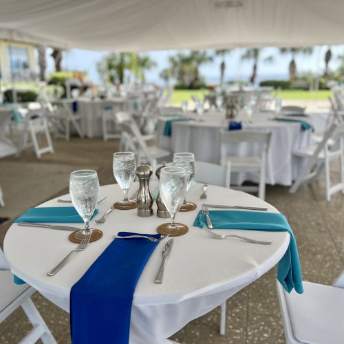 Elegant outdoor event setup under a tent with white tables, blue accents, and neatly arranged chairs.