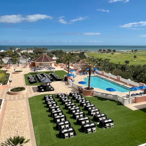 A seaside resort with a pool, outdoor dining setup, palm trees, and a golf course, located next to the ocean.