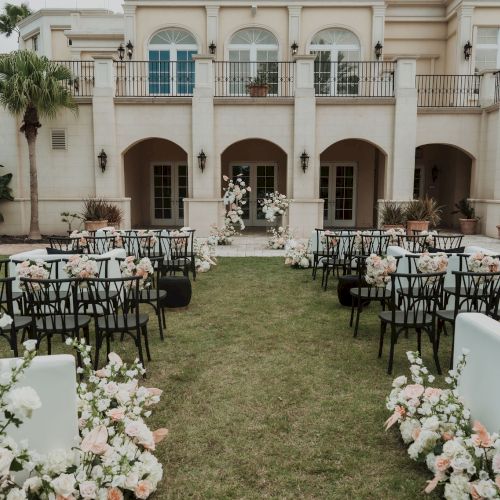 An elegant outdoor wedding setup with floral decorations, black chairs, and a champagne tower against a large beige building.