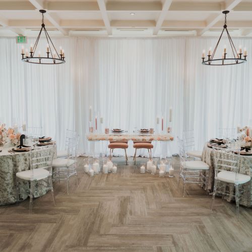 Elegant event setup with two decorated tables, transparent chairs, candles, floral arrangements, and chandeliers, in a bright room.