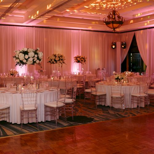 The image shows an elegantly decorated event hall with round tables, white linens, floral centerpieces, and soft pink lighting.