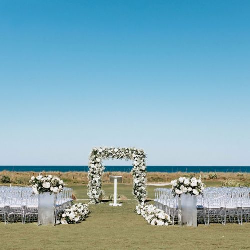 Outdoor wedding ceremony setup with white flowers, clear chairs, and an ocean backdrop under a clear blue sky.