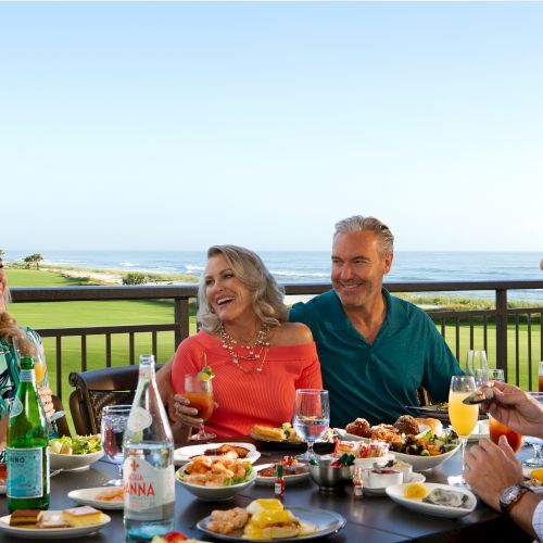 Four people enjoy a meal with drinks on a balcony overlooking the ocean and a golf course.