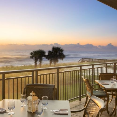 A coastal restaurant terrace at sunrise with tables set for dining; palm trees and ocean waves are in the background.