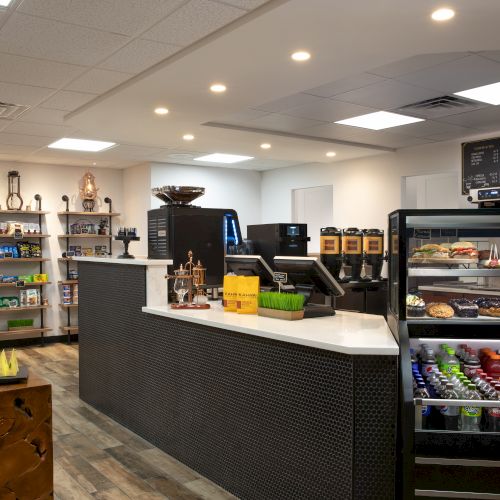 A modern café with a counter, pastry display, beverage fridge, and shelves of packaged goods. Cozy and inviting atmosphere.