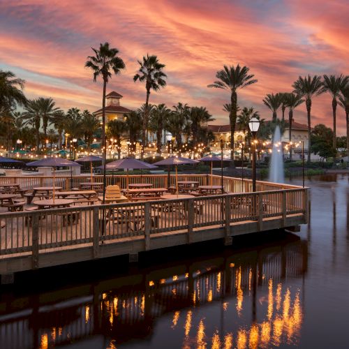 A scenic evening view of a waterfront area with palm trees, illuminated lights, and seating, under a vibrant sunset sky.