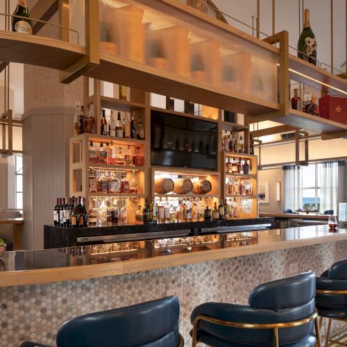 The image shows an elegant bar area with bottles of alcohol, stools, a bowl of citrus fruits, and warm lighting.