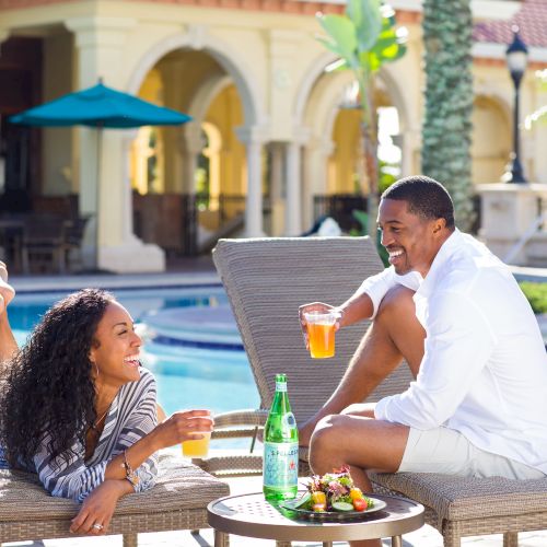 Two people relax by a pool, laughing and enjoying drinks, with a bottle of sparkling water and snacks on a table between them.
