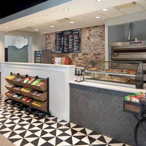 The image shows a pizzeria interior with a counter displaying pizzas, snacks, a menu on the wall, and condiment bottles on the side.