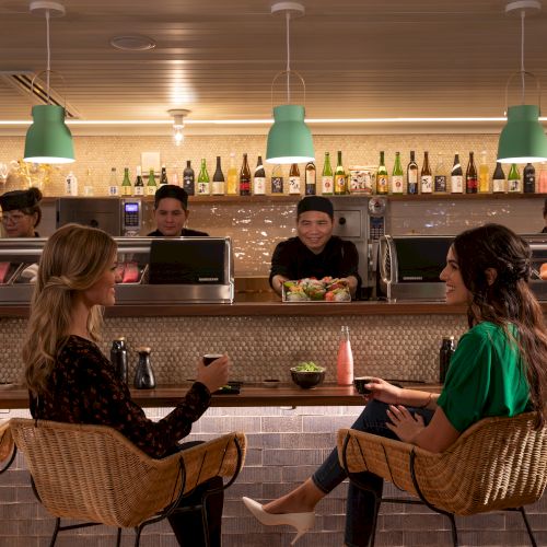 Two women sit at a sushi bar, enjoying drinks. Chefs prepare sushi behind the counter, with bottles displayed on shelves.