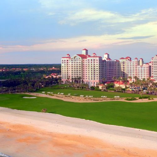 A beachside resort with a large hotel, golf course, and palm trees, set against a pastel sky along a sandy coastline.