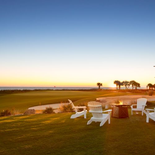 A sunset view with chairs around a fire pit on a grassy area, near a beachside house and palm trees.