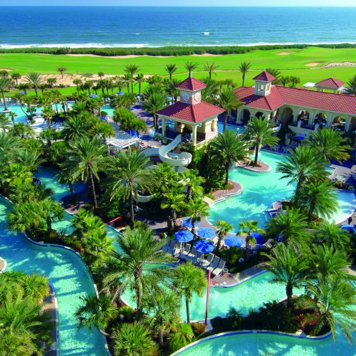 A tropical resort with pools, palm trees, and a red-roofed pavilion, set against a backdrop of ocean and greenery.