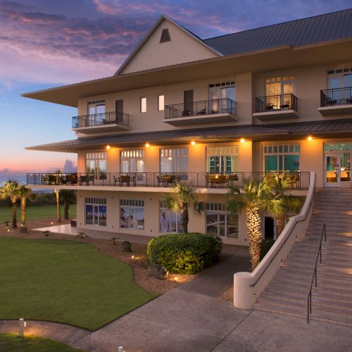 A large building with balconies and warm lights at sunset, surrounded by palm trees and a well-kept lawn, overlooks the ocean.