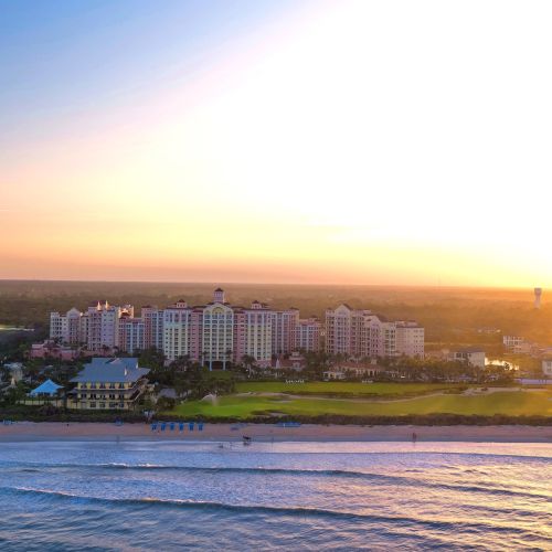 A beachfront resort with several tall buildings and a golf course, set against a bright, glowing sunset sky.