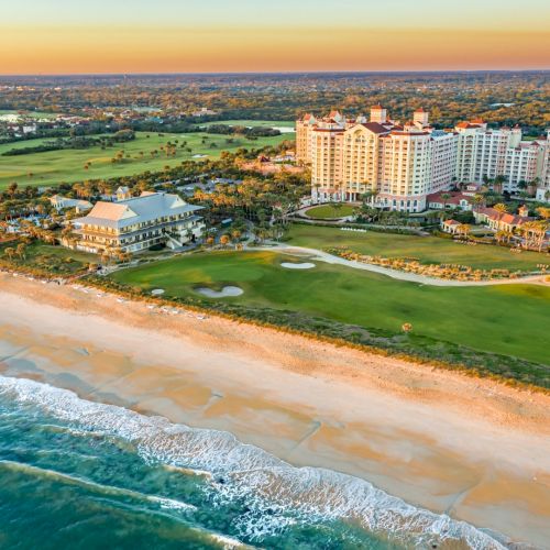 Aerial view of a beachfront resort with a sandy beach, golf course, and lush greenery, set against a coastal landscape at sunset.