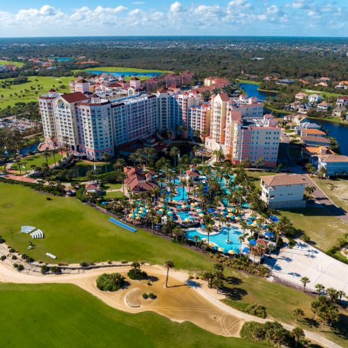 Aerial view of a large resort with pools, surrounded by greenery, golf course, and nearby residential area.