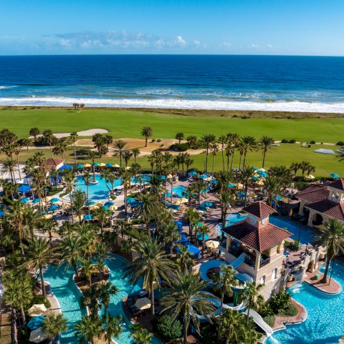 Aerial view of a resort with pools, palm trees, and golf course by the ocean. Bright blue umbrellas and chairs around the pool area.