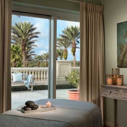 A serene room with a massage table, lit candles, artwork, and window view of palm trees and a patio area.