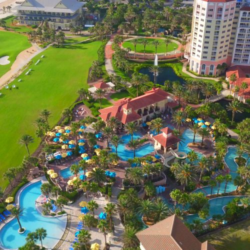 Aerial view of a coastal resort with a golf course, pool area, palm trees, and buildings near the beach, featuring umbrellas and green lawns.