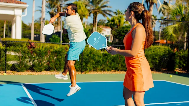 Two players volley on an outdoor pickleball court with paddles and a sunny blue sky behind them.