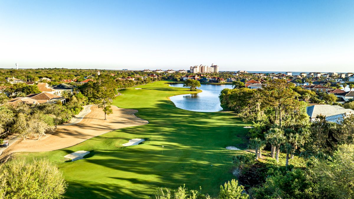 Aerial view of a golf course with a sand trap, green, and pond, surrounded by trees, houses, and distant buildings under a clear blue sky.