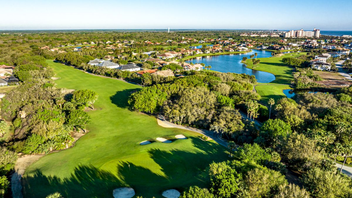 Aerial view of a lush golf course with sand traps, surrounded by trees, homes, and a winding waterway under a clear blue sky.