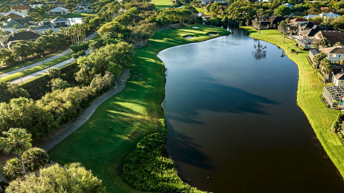 Aerial view of a golf course, with a large pond, lush greenery, and nearby houses under a clear sky.
