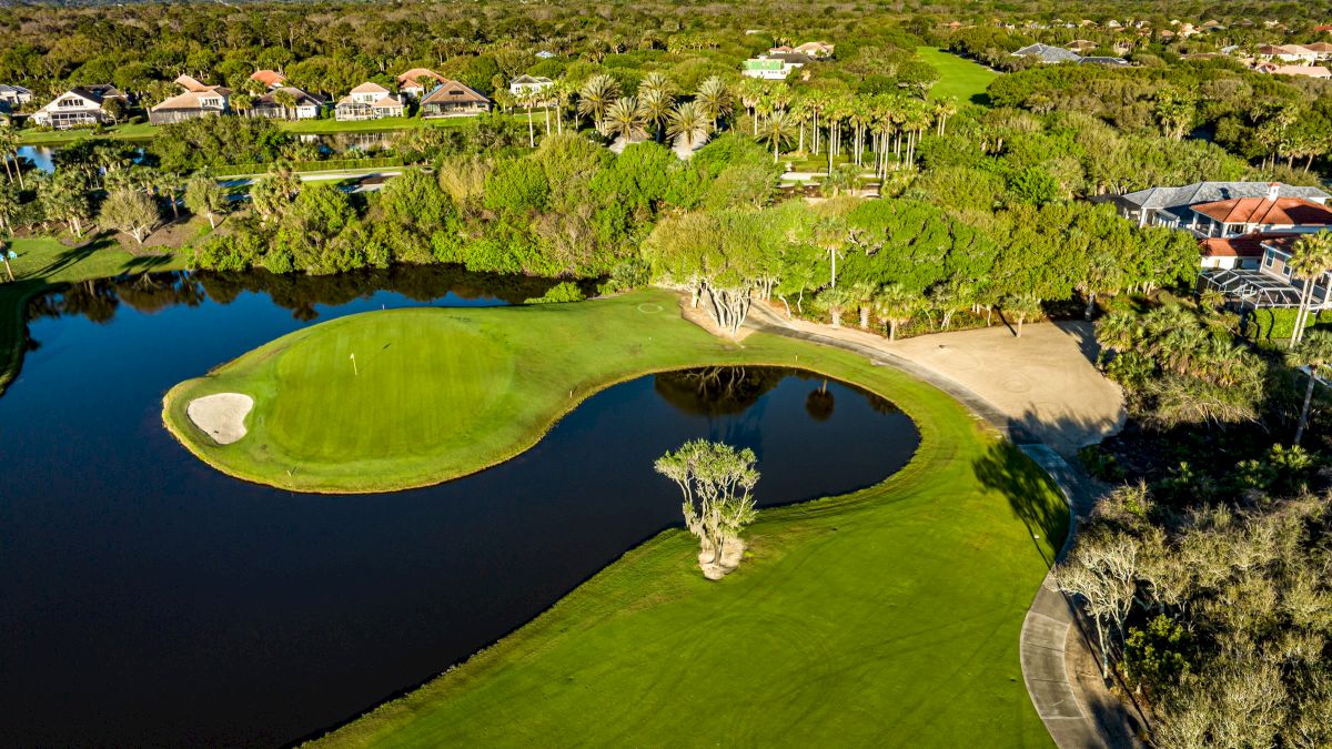 Aerial view of a lush golf course with a green surrounded by water, trees, and nearby houses.