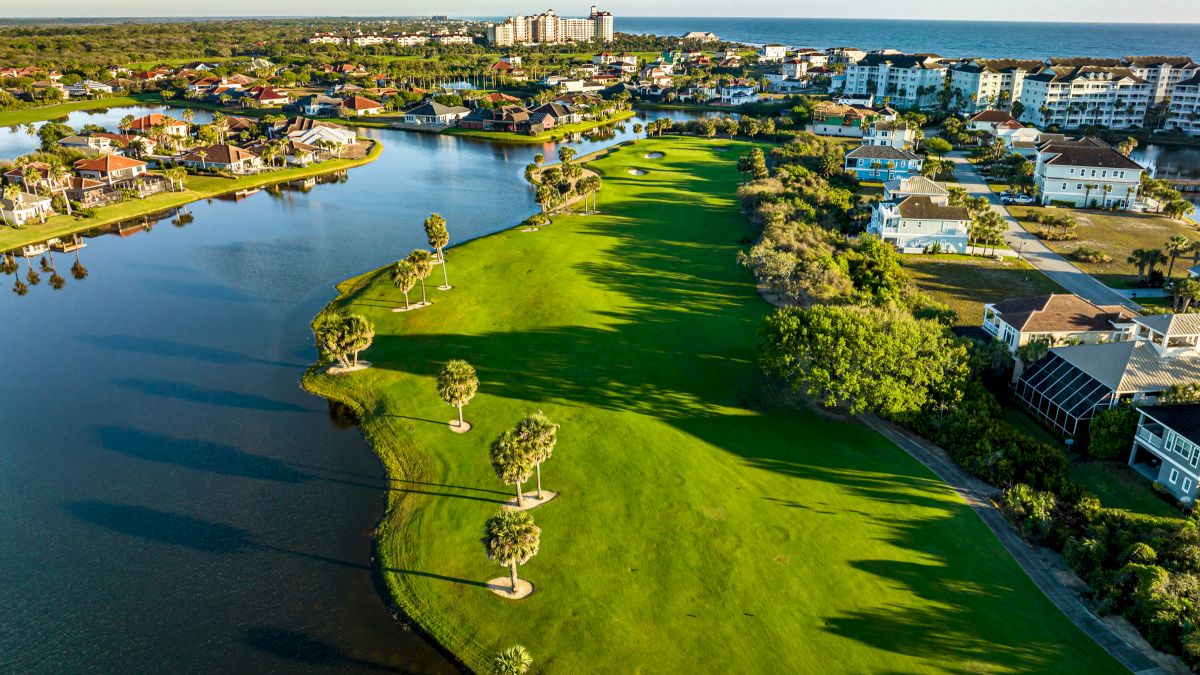 Aerial view of a waterfront landscape with green lawns, palm trees, a lake, and houses near the ocean in the background.