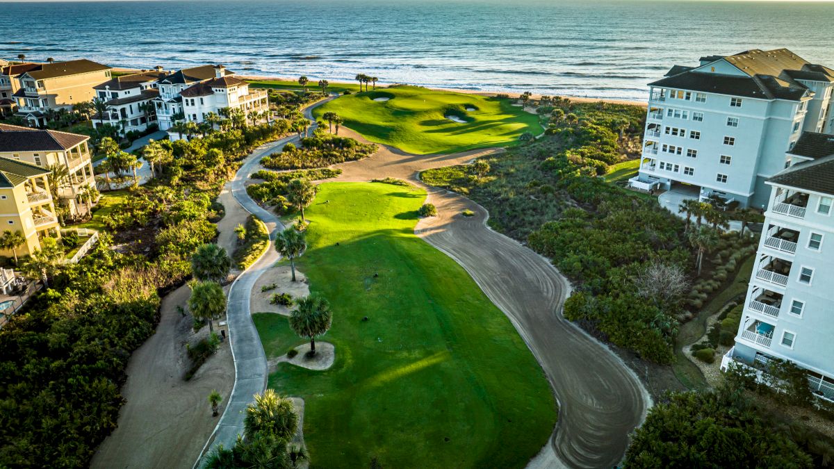 Aerial view of a coastal golf course with nearby residential buildings and a path by the ocean, bordered by lush greenery.
