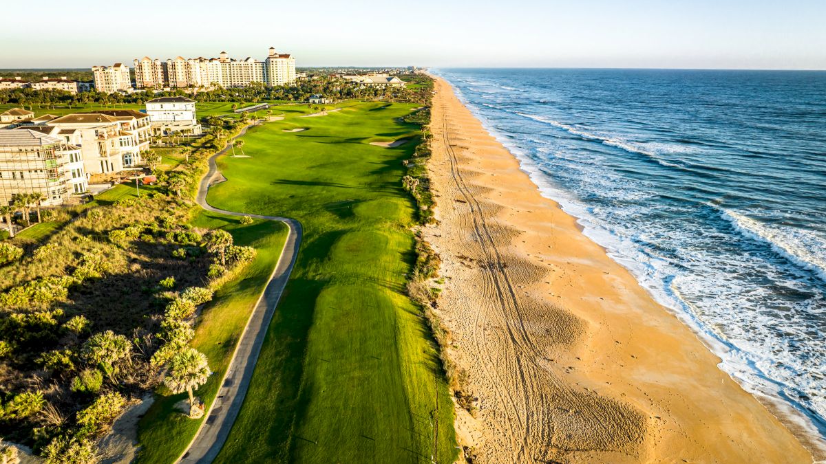 Aerial view of a beachfront area with buildings, golf course, and shoreline with waves.