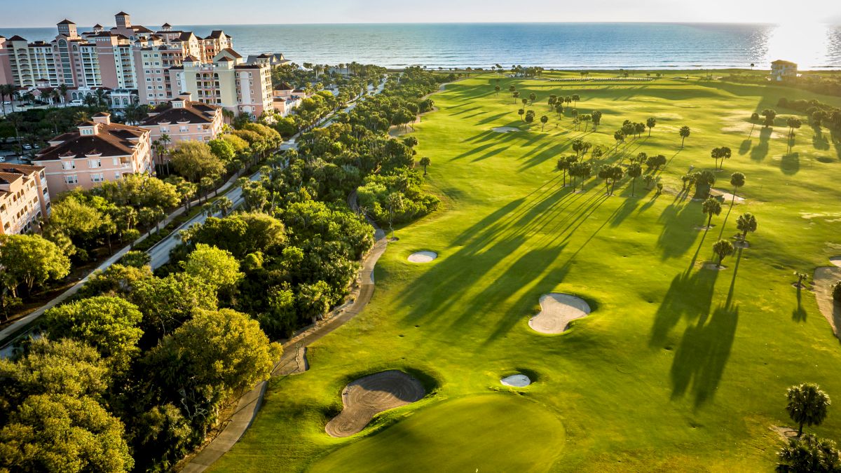 A coastal golf course with adjacent buildings, trees, and a view of the ocean at sunset.
