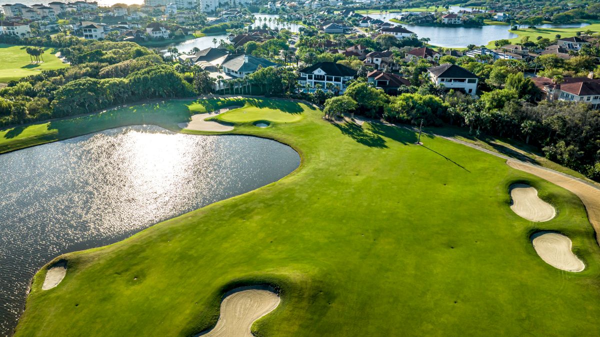 Aerial view of a golf course with sand bunkers, a large water feature, and houses in the background near a coastal area.
