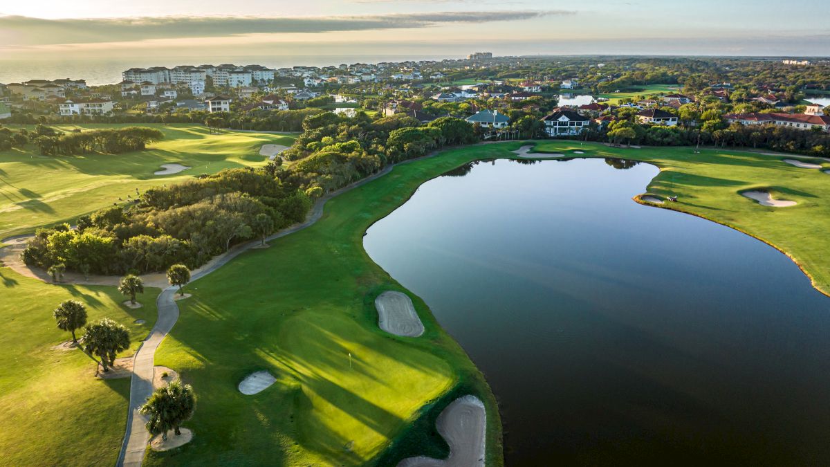 An aerial view of a golf course with sand traps, a pond, and surrounding residential buildings under a blue sky at sunrise or sunset.