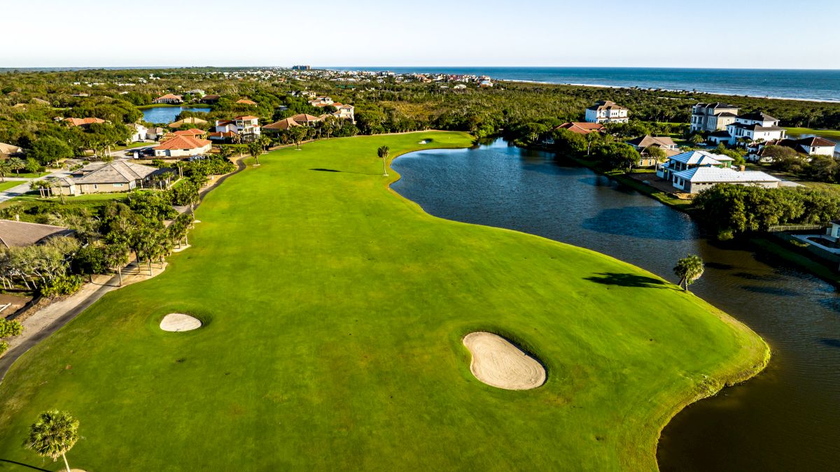 Aerial view of a golf course with sand traps, houses, water features, and a distant coastline under a clear blue sky.