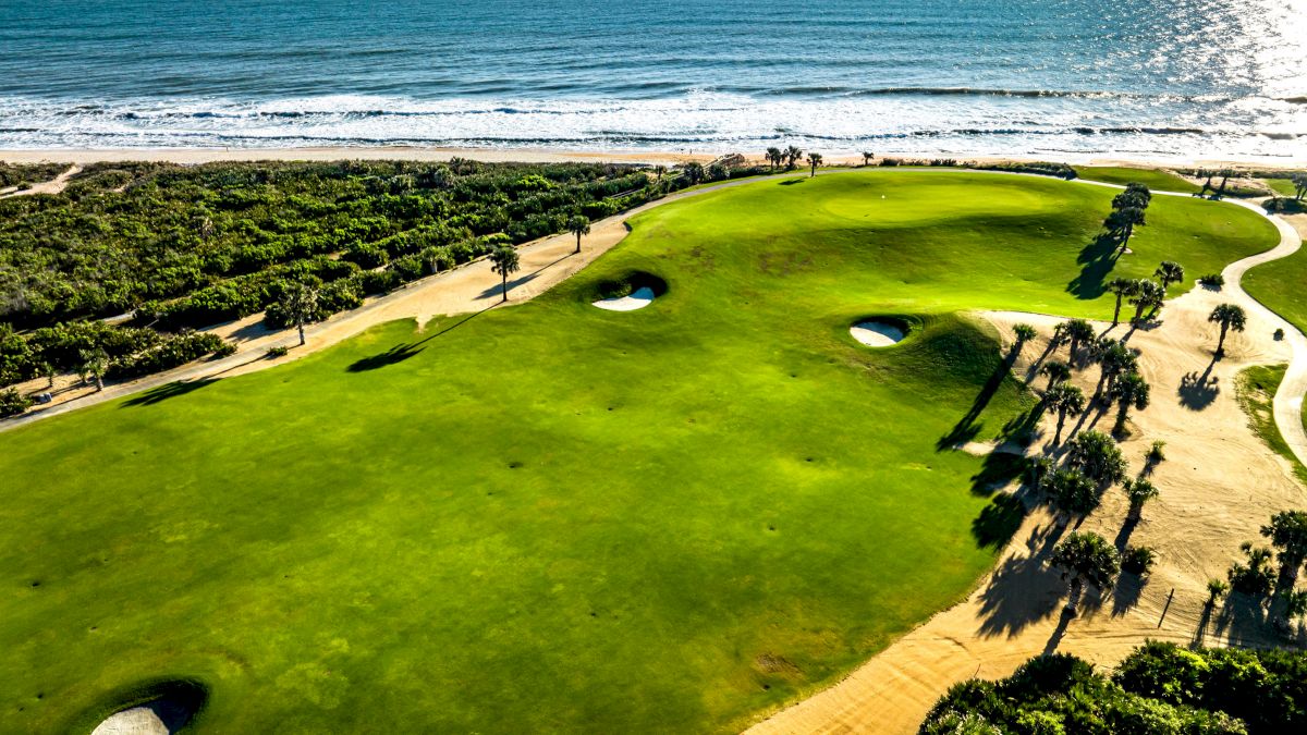 A lush golf course with sand bunkers, bordered by the ocean and surrounded by greenery, under a clear blue sky.