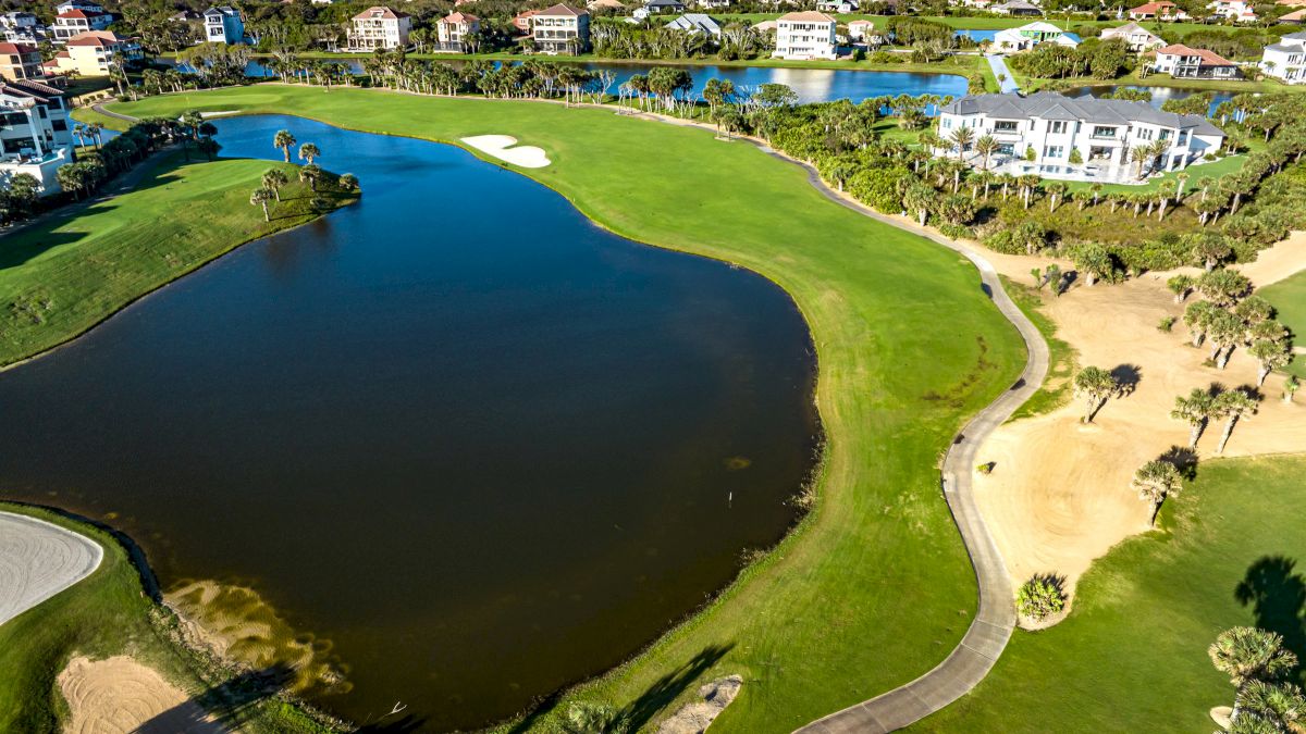 The image shows a golf course with a water hazard, sand traps, a winding path, surrounded by residential buildings and greenery.