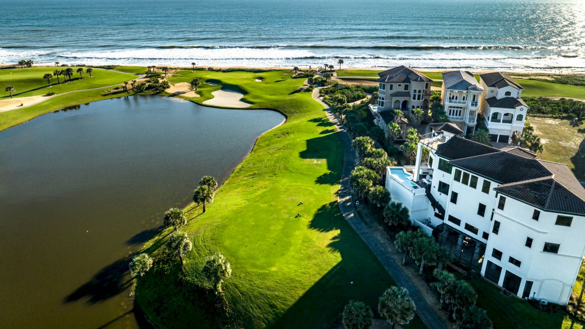 Aerial view of a scenic seaside golf course with lush greens, a water hazard, luxury homes, and an ocean backdrop under clear skies.
