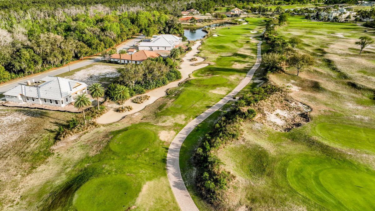 The image shows a scenic aerial view of a golf course with lush greenery, a winding path, and houses nearby.