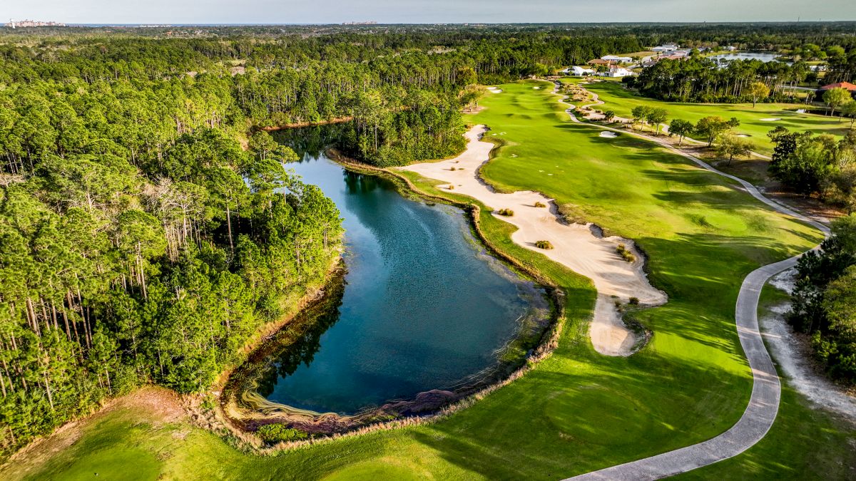 Aerial view of a golf course with a lake, surrounded by trees, green fairways, sandy bunkers, and a winding path.
